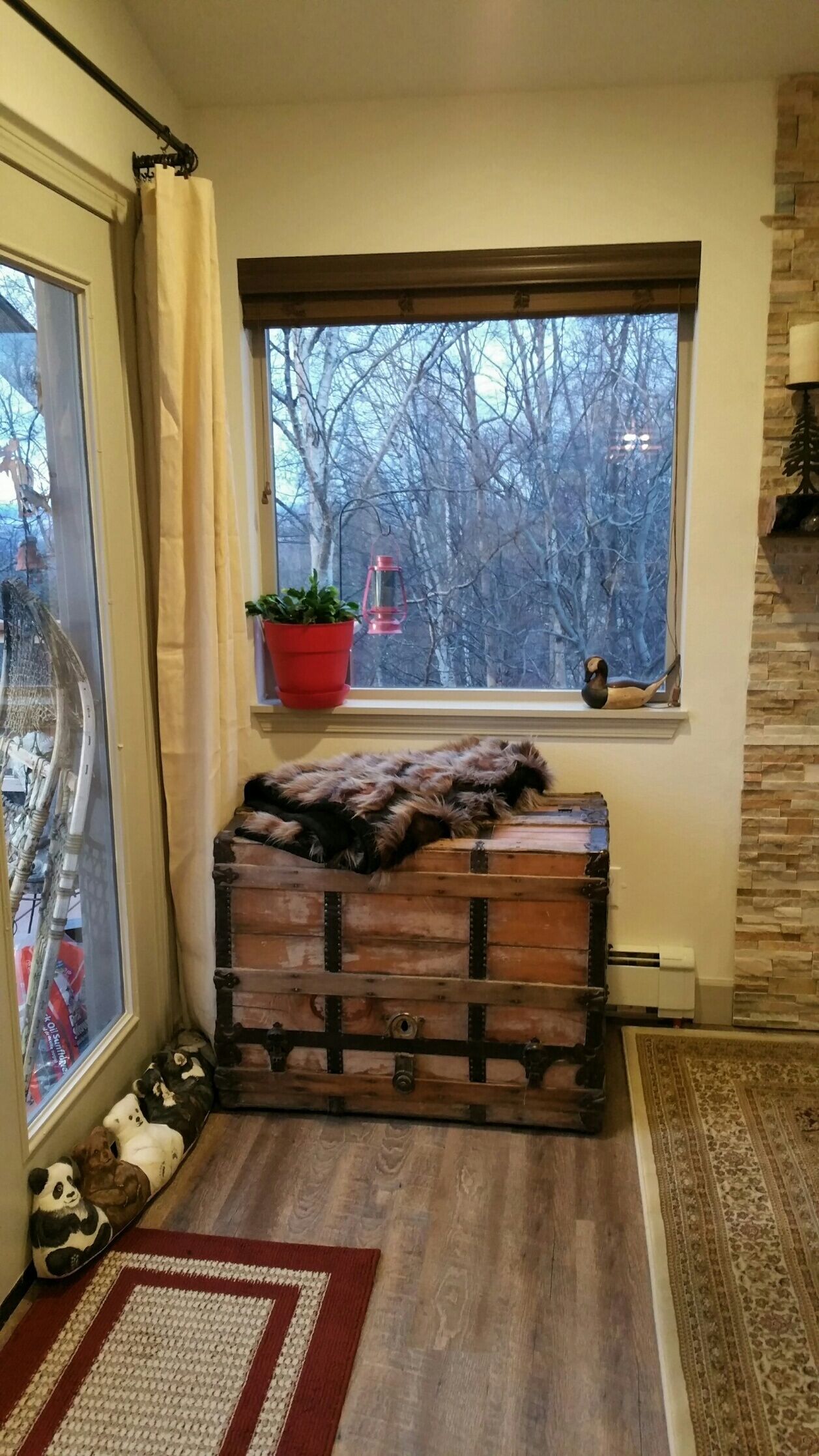Un coin douillet avec un coffre en bois, une fenêtre donnant sur des arbres, une plante en pot rouge et un objet décoratif en forme de panda.