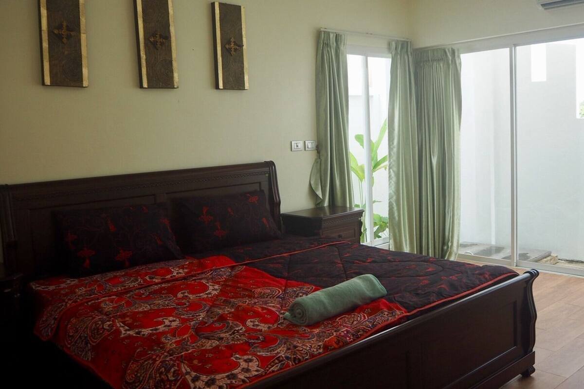 A bedroom with a large bed, a nightstand, a window with curtains, and a view of the outside.
