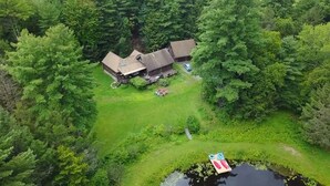 Une vue aérienne d’un chalet rustique entouré d’une forêt dense, avec un petit quai qui s’avance dans un étang.