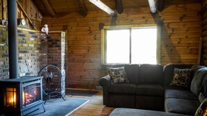 L’intérieur d’un chalet accueillant, avec un poêle à bois, une cheminée en briques, un canapé et une grande fenêtre.