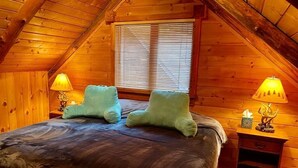 Une chambre confortable située dans les combles, avec un plafond en bois, un lit avec deux oreillers, une table de chevet avec une lampe et une fenêtre équipée de stores.