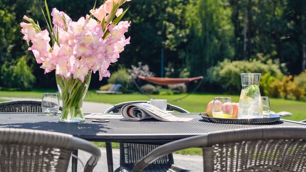 Una mesa de comedor al aire libre con un jarrón de flores rosas, un periódico y una bandeja con fruta y bebidas.
