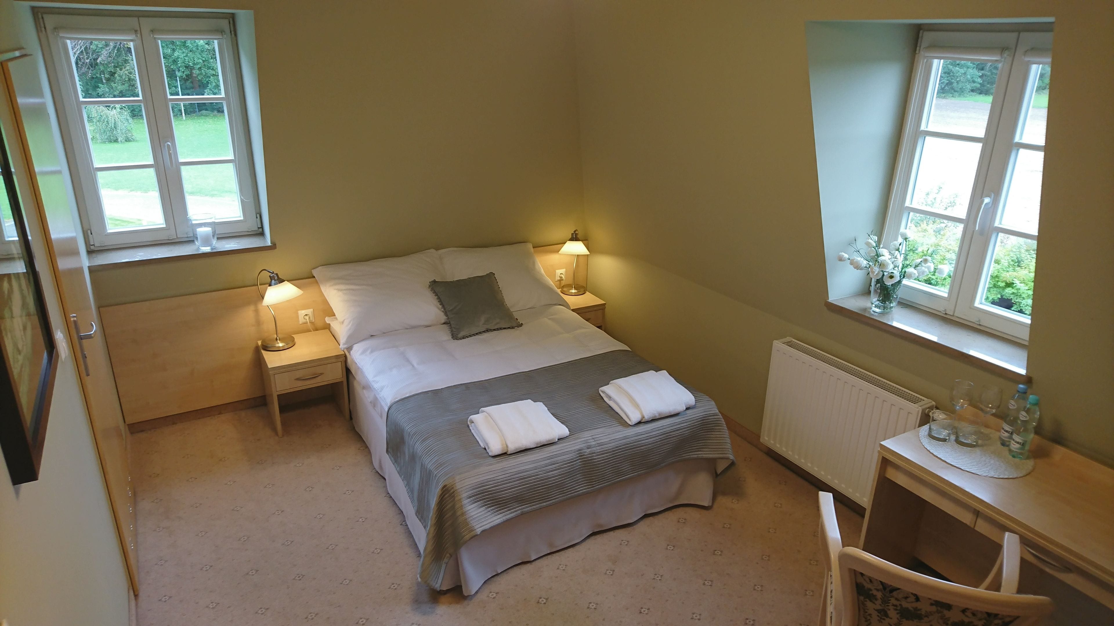 A bedroom with a bed, two bedside lamps, a nightstand, a chair, a small table with a vase, and two windows.