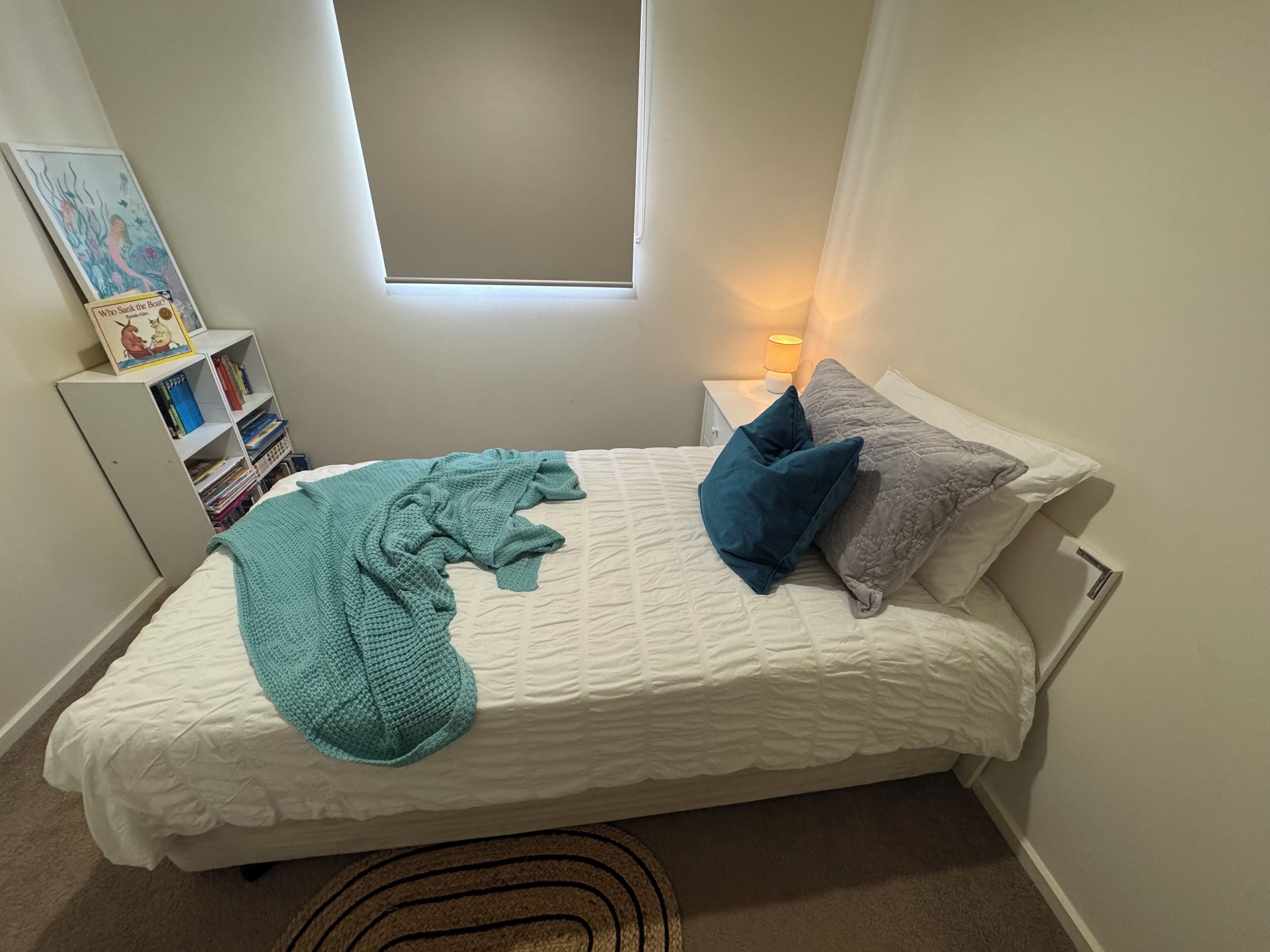 A bedroom with a bed, pillows, a knitted blanket, a bookshelf, and a window.