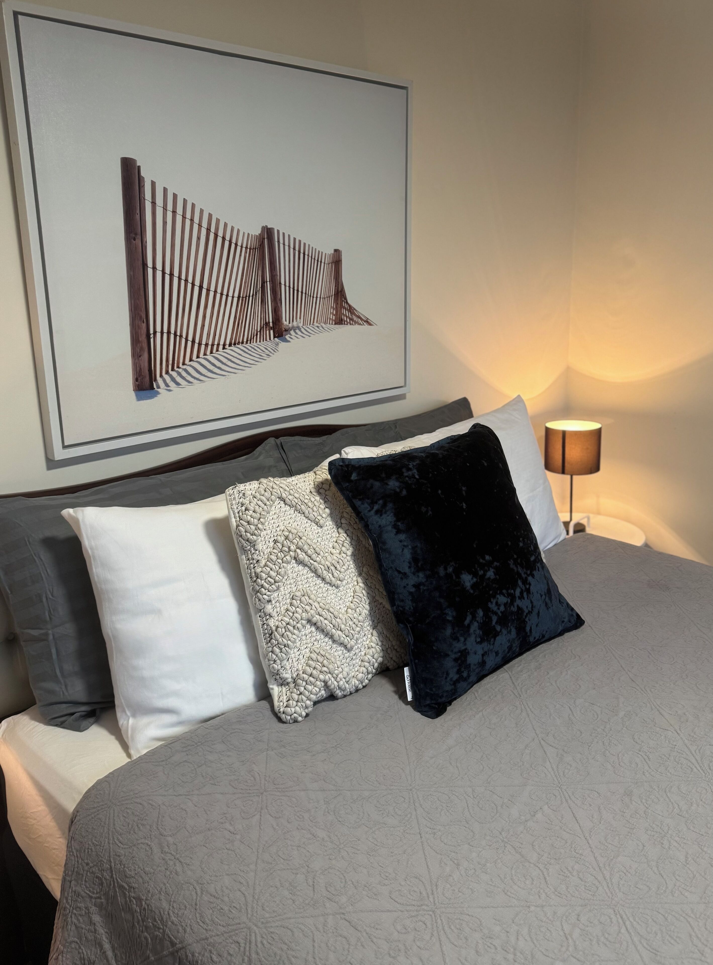A neatly made bed with a grey bedspread and three pillows, a framed artwork above the bed, and a bedside lamp.