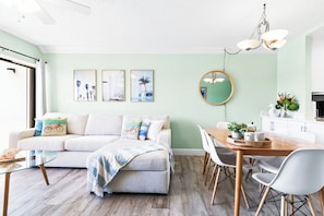 A modern living room with a sofa, coffee table, dining table, chairs, and a kitchen area with cabinets and a hanging light fixture.