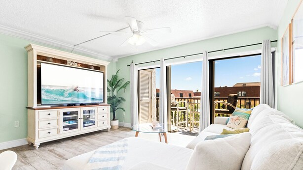 A living room with a TV, sofa, and a balcony with outdoor furniture. - Perfect Family Getaway Steps from Cocoa Beach Pier with Oceanfront Heated Pool (Cocoa Beach)