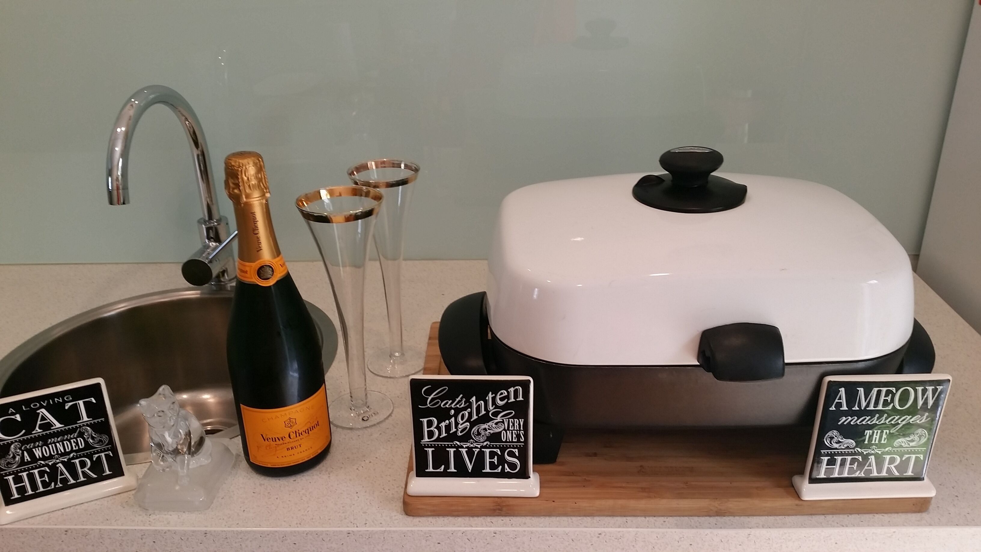A kitchen counter with a bottle of Veuve Clicquot champagne, two glasses, a toaster, and signs with cat-themed messages.