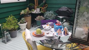 Mesa de vidrio dispuesta con platos, tazas y objetos decorativos en un balcón con plantas en macetas y una pared verde.