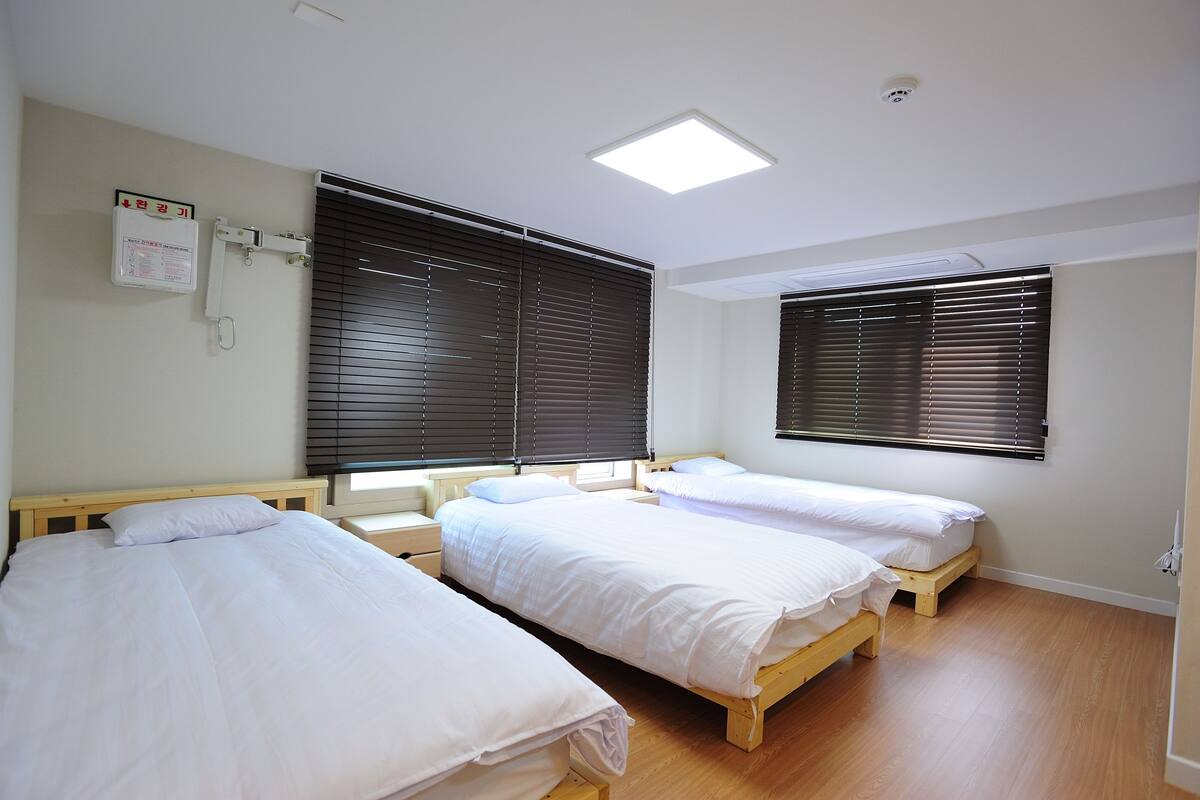 Habitación con tres camas individuales, piso de madera y ventanas amplias con persianas.