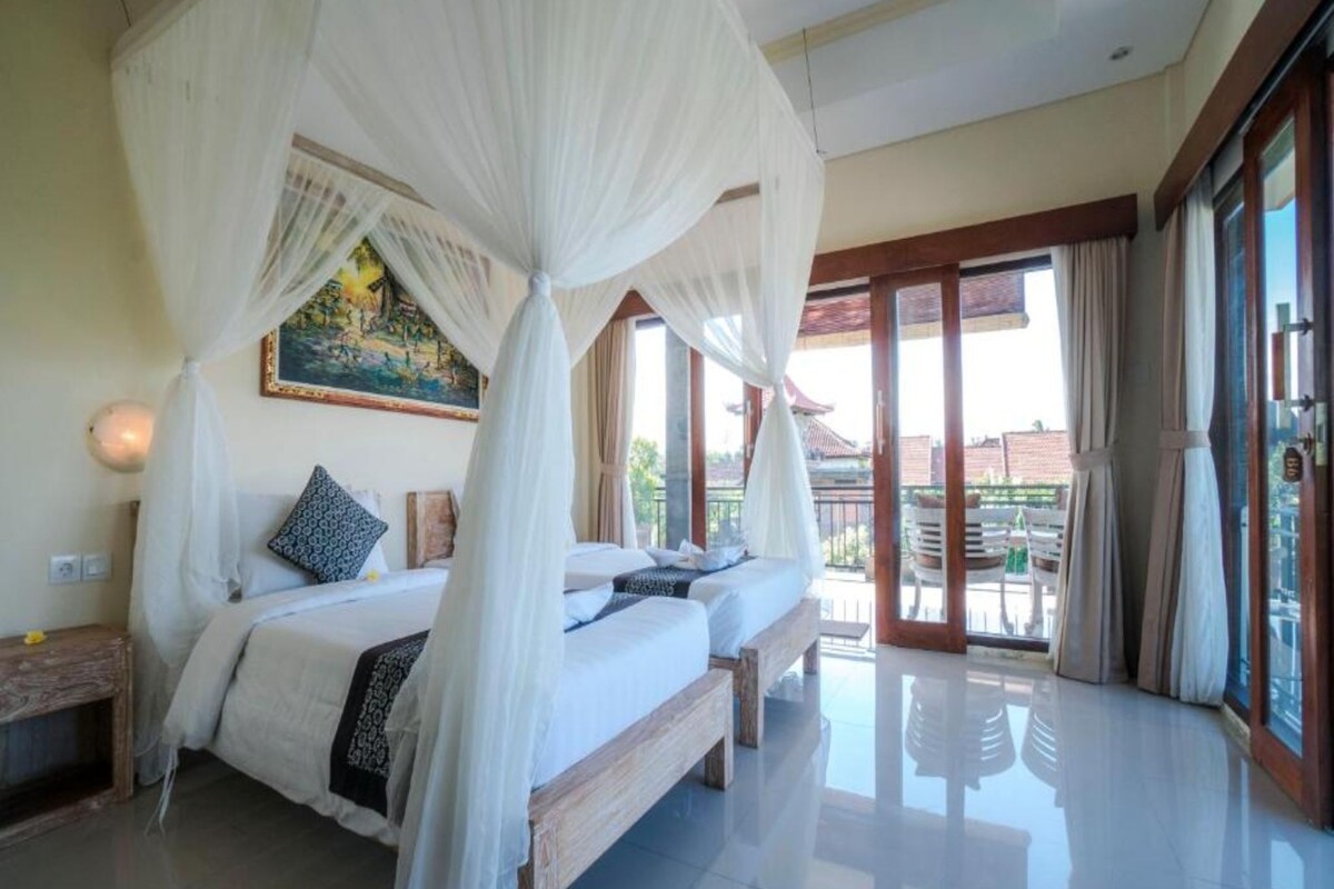 A bedroom with a canopy bed, a painting on the wall, and a view of the outside through large windows.