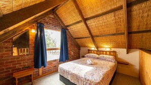 A bedroom with a sloped ceiling, a brick wall, a wooden bench, a bed with a patterned bedspread, and a window with curtains. - Grampians Pioneer stone cabin (Halls Gap)