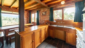 A rustic kitchen with wooden cabinets, a large window with curtains, and a stone-tiled floor. - Grampians Pioneer stone cabin (Halls Gap)