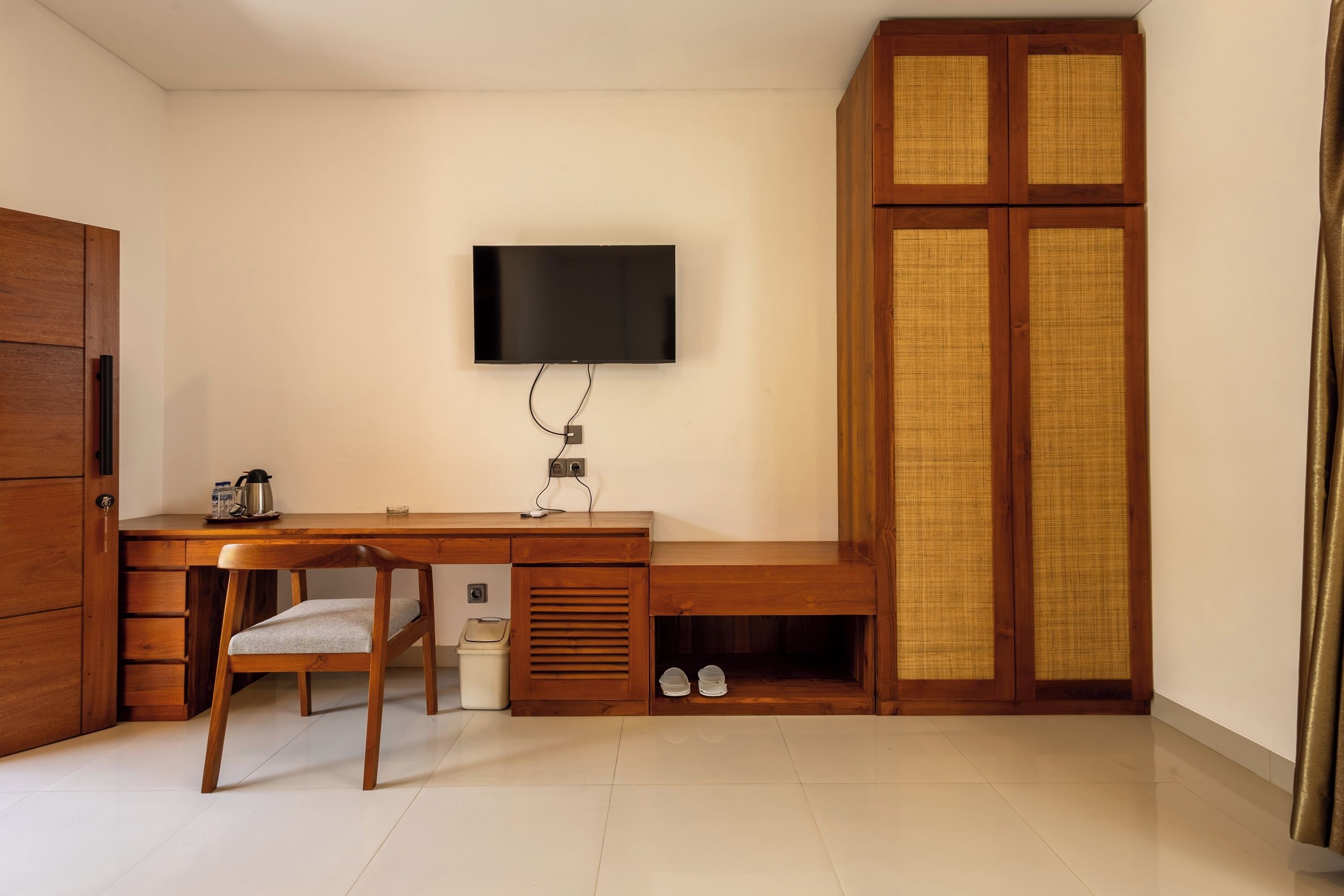 A room with a wooden desk, a chair, a television mounted on the wall, and a wardrobe with a woven door.