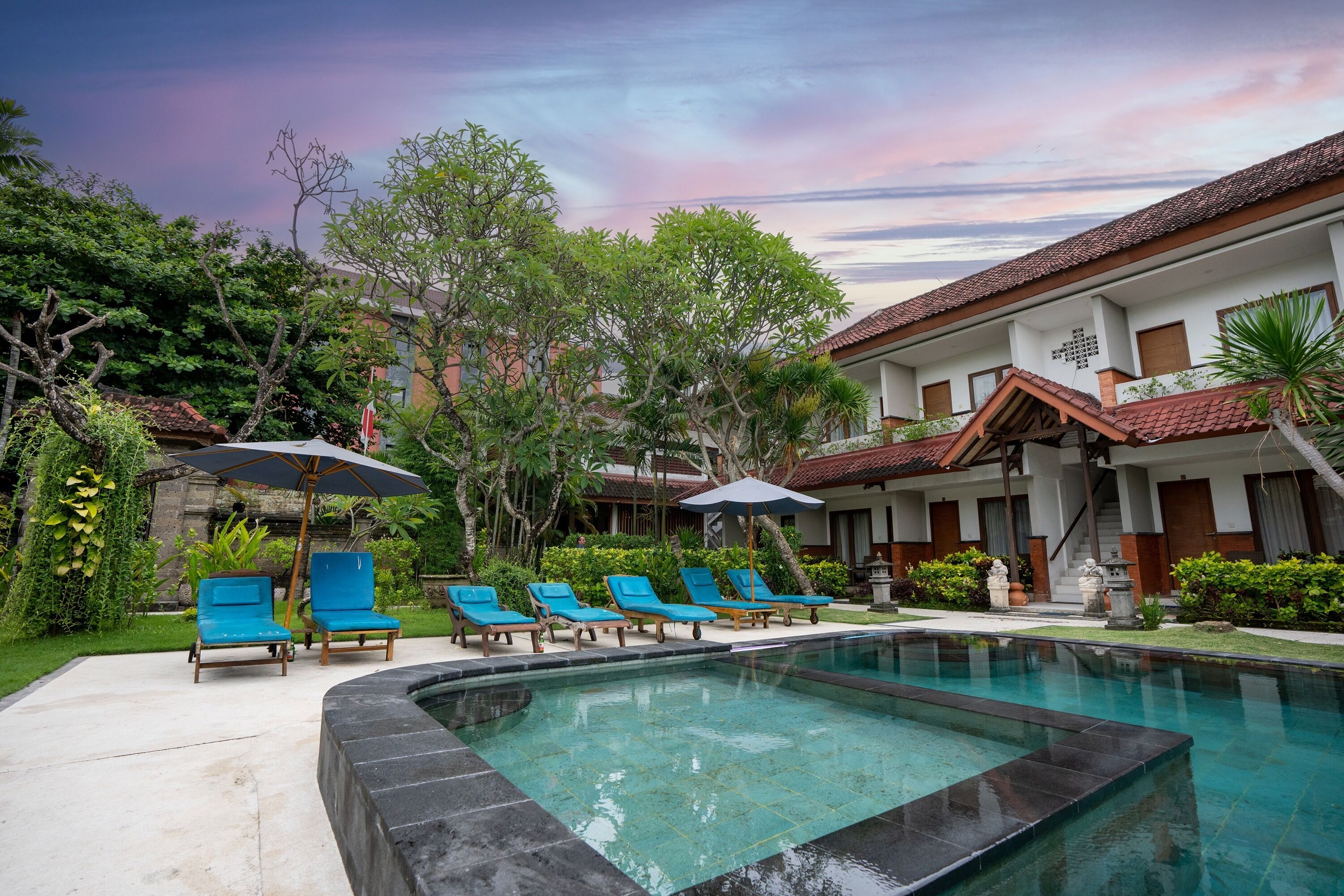 A swimming pool with lounge chairs and umbrellas, surrounded by greenery and a building with a tiled roof.