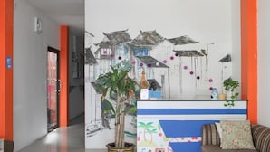 Un espace d’accueil agrémenté d’une fresque murale abstraite aux couleurs vives, d’une plante en pot et d’un canapé avec un coussin à motifs.