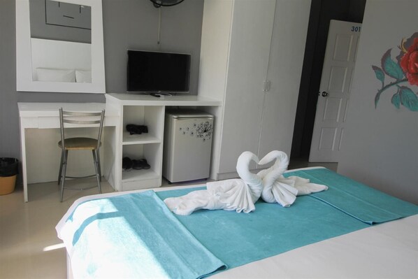 A hotel room with a bed, a desk with a television, a refrigerator, a chair, and a door with a number. - Guan Chao Yin Hotel (Patong)