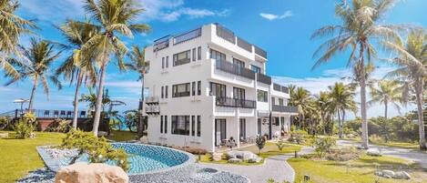 A modern multi-story hotel with a pool, surrounded by palm trees and a clear sky.