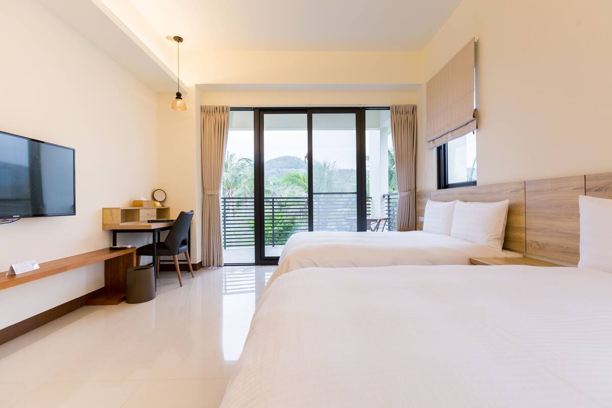 A hotel room with a large bed, a desk with a chair, a television, and a balcony with curtains.