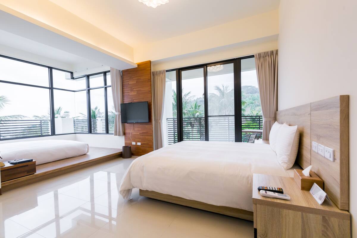 A modern hotel room with a large bed, a smaller bed, a TV, and a balcony with a view of palm trees.