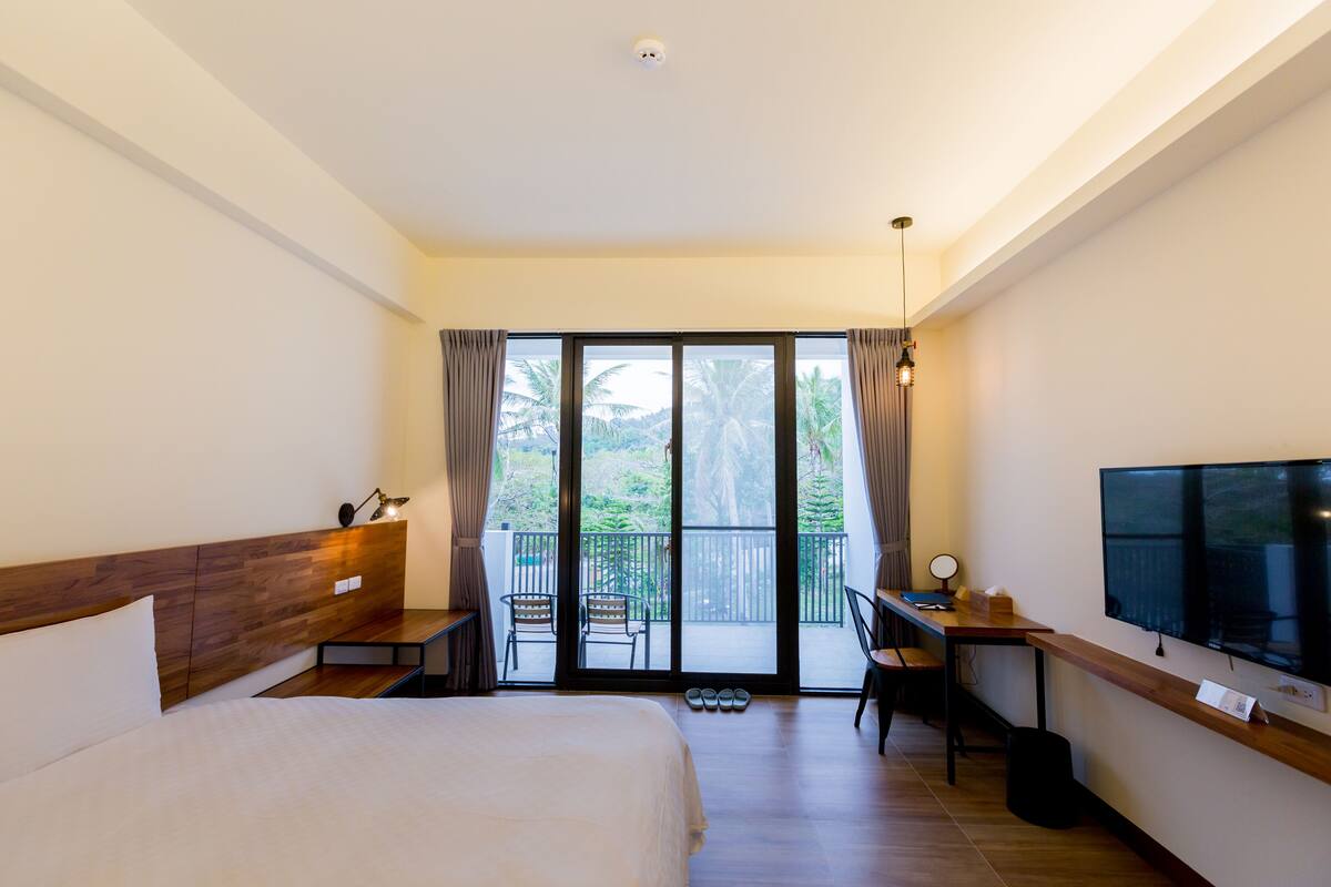 A hotel room with a bed, a desk with a chair, a television, and a balcony with a view of palm trees.