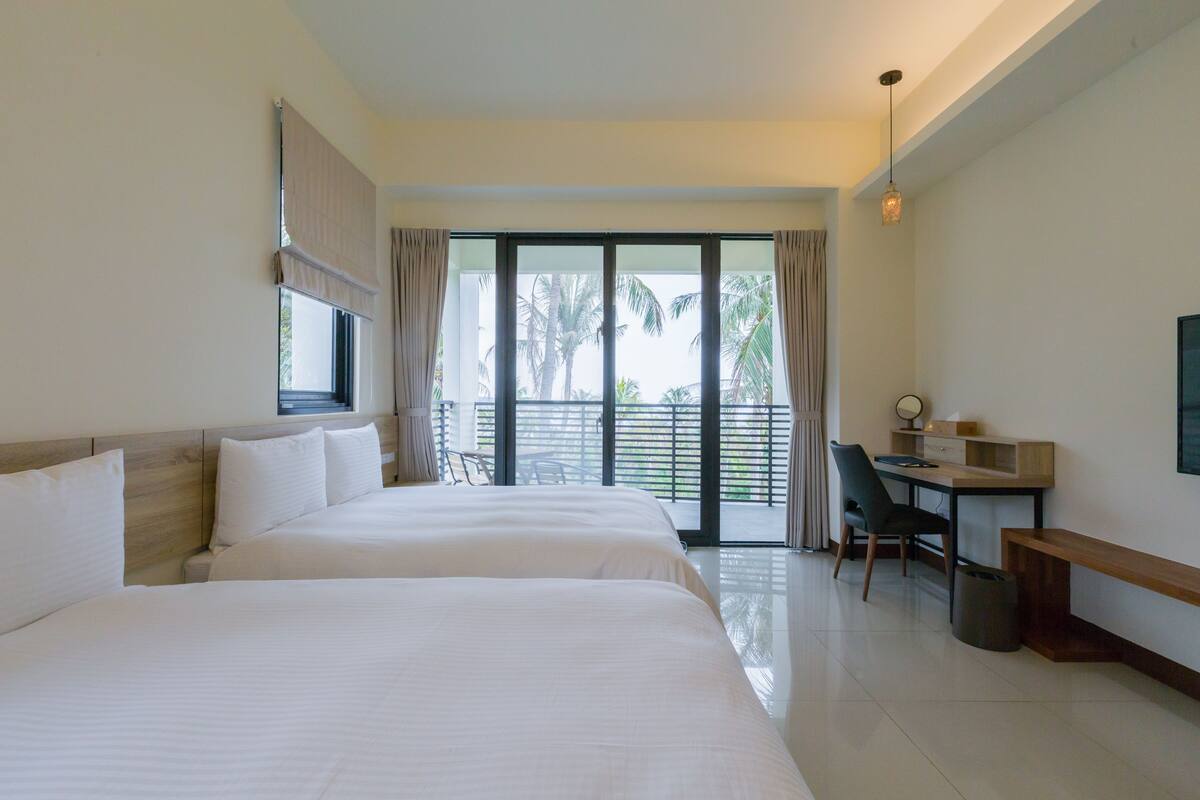 A hotel room with two beds, a desk, a chair, a bench, and a view of palm trees through the glass door.