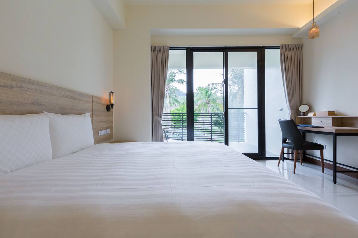 A hotel room with a large bed, a desk with a chair, and a view of palm trees through the window.