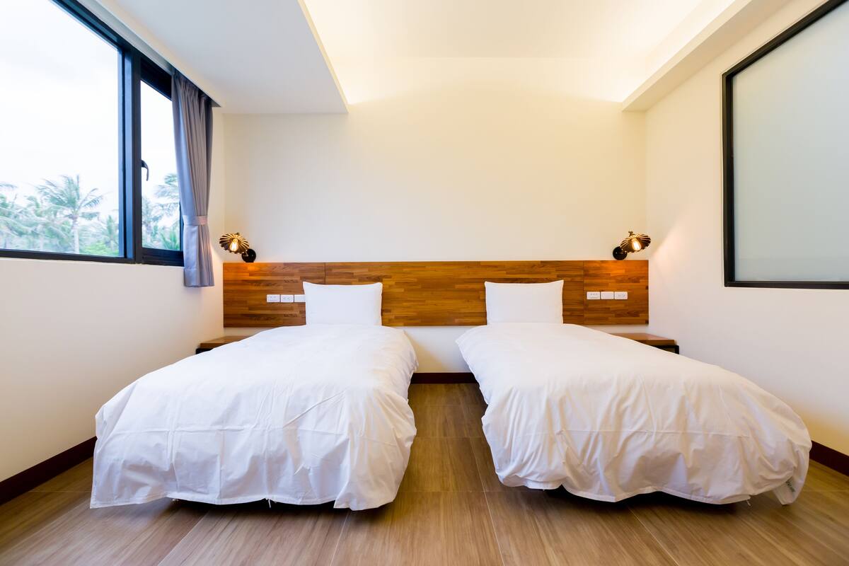 A hotel room with two beds, a wooden headboard, a large window with curtains, and a wall-mounted lamp.