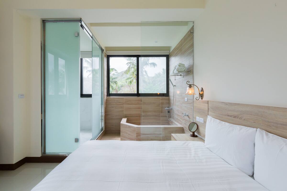 A modern hotel room with a large bed, a glass door leading to a balcony, a built-in bathtub, and a view of palm trees.