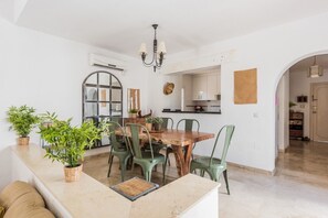 Un comedor con una mesa de madera y sillas verdes, una cocina al fondo, y una decoración en tonos blancos.