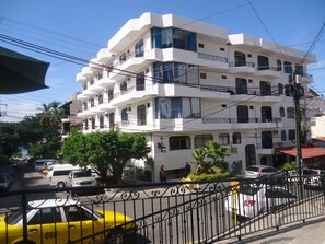 Un immeuble blanc à plusieurs étages, doté de balcons, avec un parking et un taxi jaune garé devant.