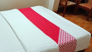 Ein ordentlich bezogenes Bett mit weißer und roter Bettdecke, einem gemusterten Kopfkissen und einem kleinen Nachttisch mit Blumen in einer Vase.