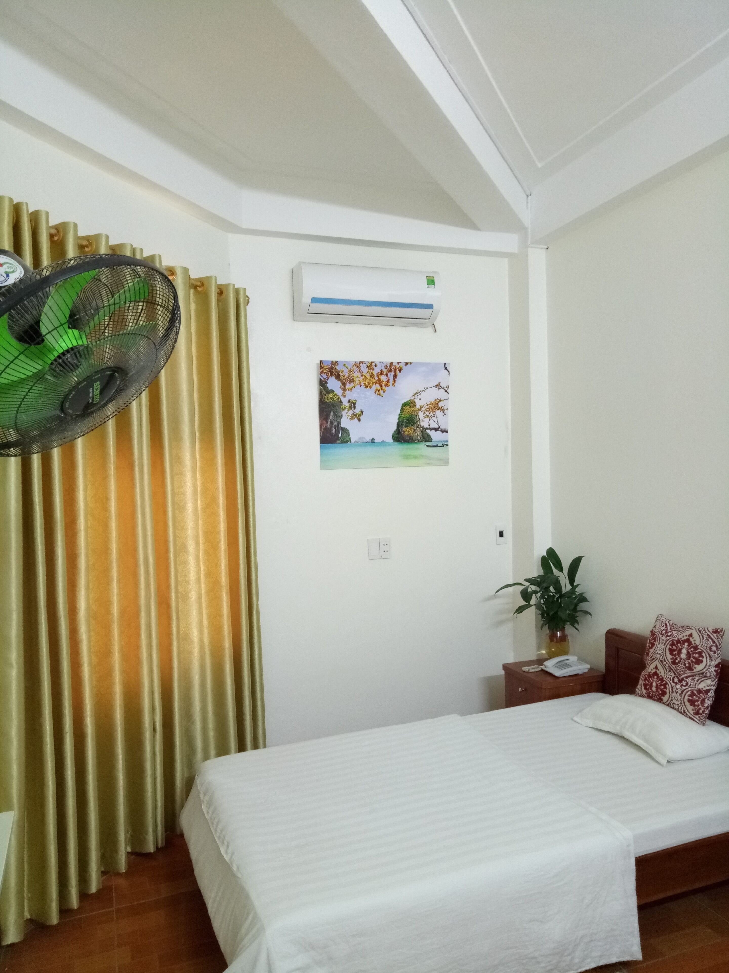 A hotel room with a bed, a nightstand, a wall-mounted air conditioner, a window with curtains, and a wall-mounted picture.