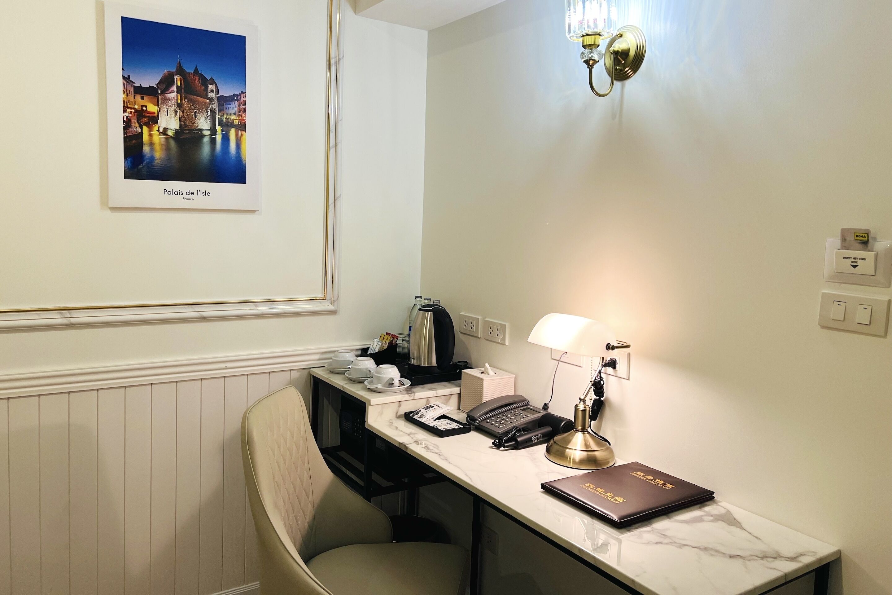 A hotel room with a marble desk, a chair, a lamp, a phone, a laptop, and a framed picture of a canal.
