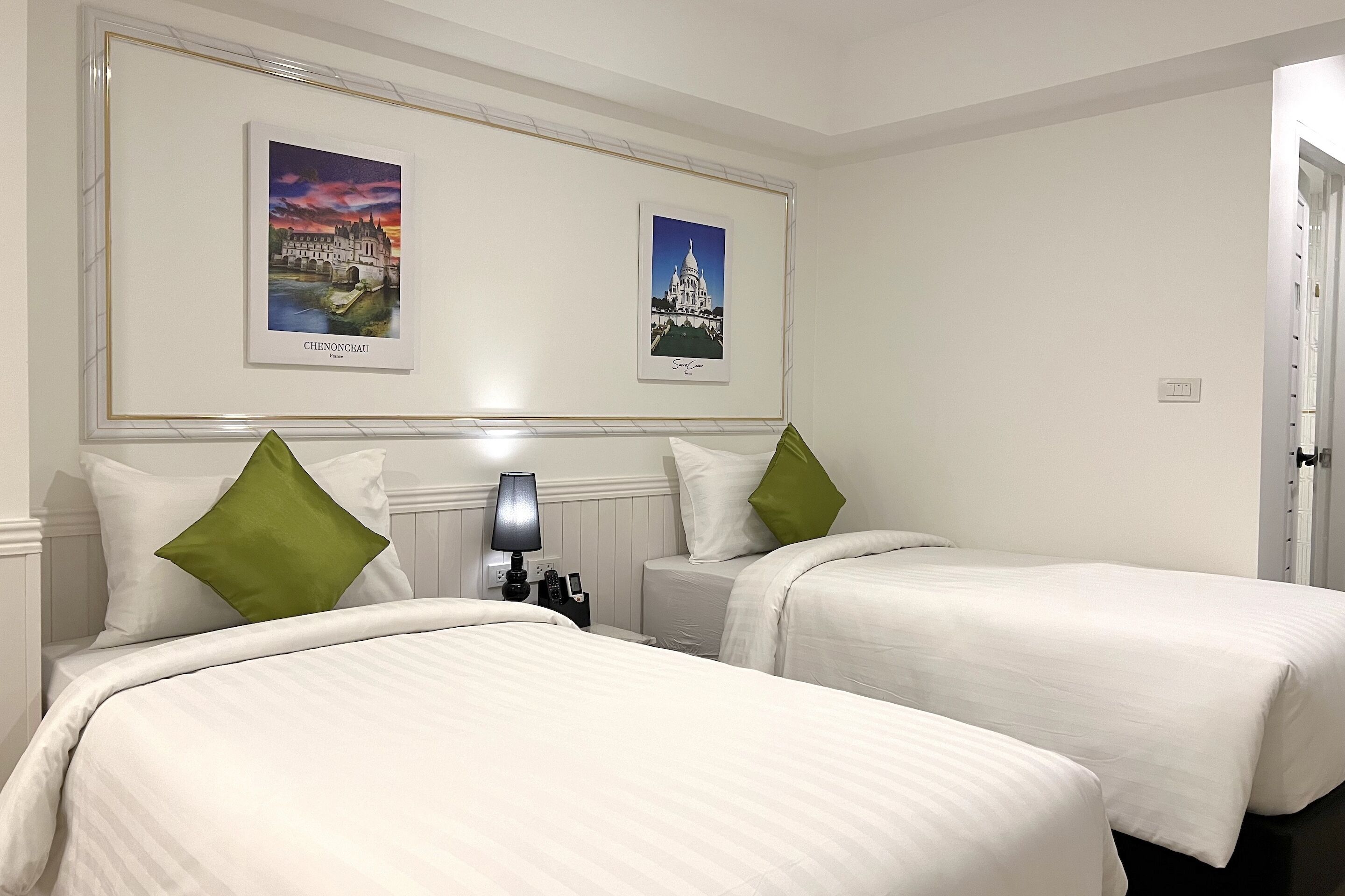 A hotel room with two beds, white bedding, green pillows, a nightstand with a lamp, and two framed pictures on the wall.