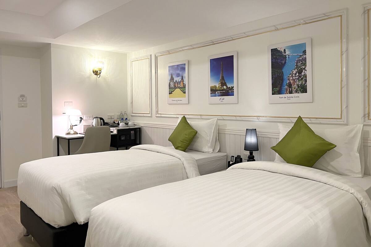 A hotel room with two beds, a desk, and a chair. There are three framed pictures on the wall.