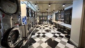 A laundry area with multiple washing machines, a checkered floor, and a seating area with tables and stools. - Travelier Hostel (Bangkok)