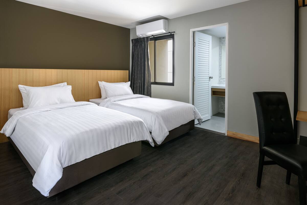 A hotel room with two beds, a wooden headboard, a window with curtains, a door leading to a bathroom, and a black chair.