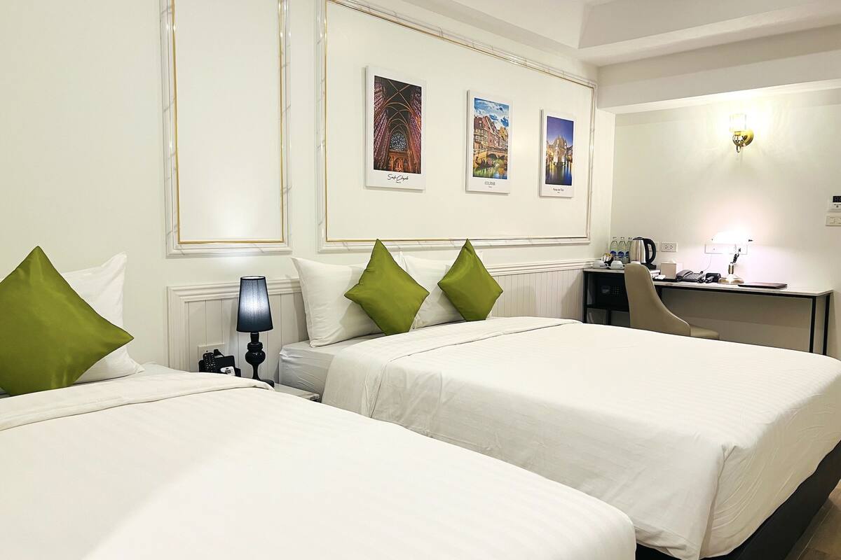 A hotel room with two beds, a desk, a chair, and a lamp. There are three framed pictures on the wall.