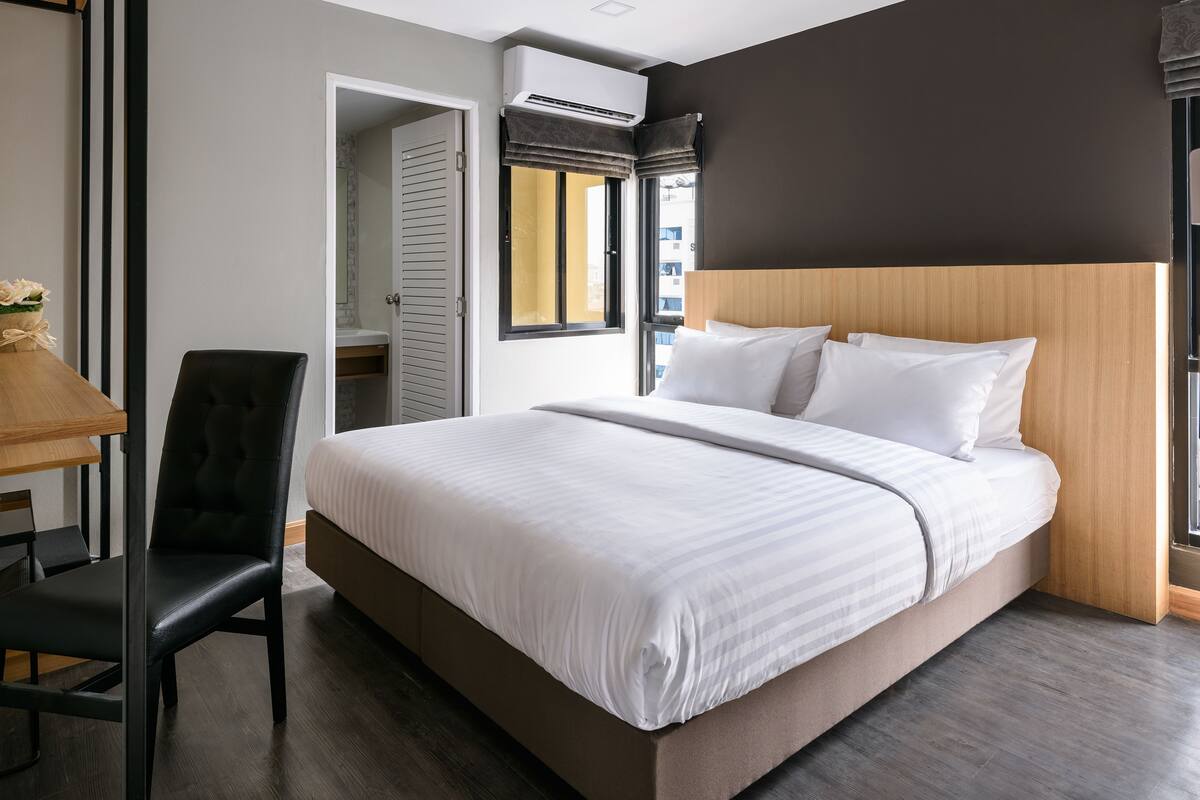A modern hotel room with a large bed, a wooden headboard, a desk with a chair, and a window with blinds.