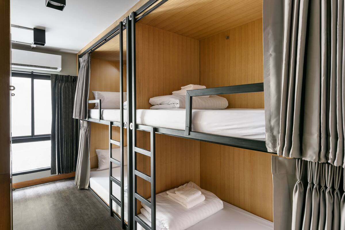 A bunk bed room with wooden paneling, a window with curtains, and a ceiling-mounted air conditioning unit.