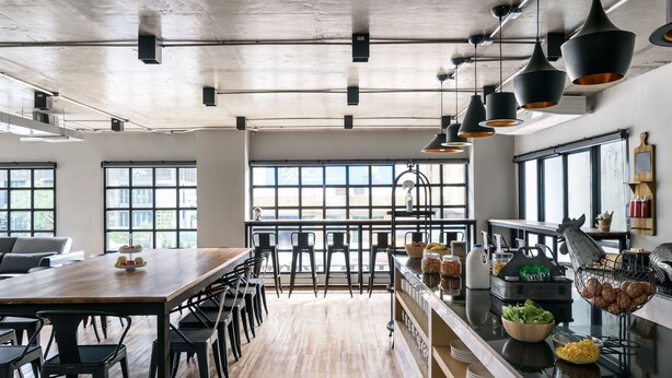 A spacious kitchen with a long wooden table, bar stools, and modern pendant lighting. - Travelier Hostel (Bangkok)