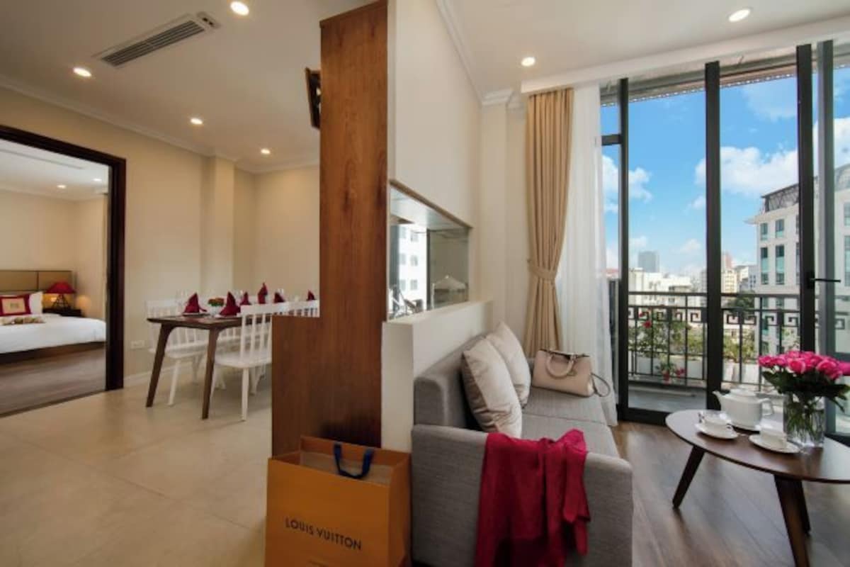 Une chambre d’hôtel comprenant un lit, une table à manger avec des chaises, un canapé, une petite table avec un vase de fleurs et une grande fenêtre donnant sur la ville.