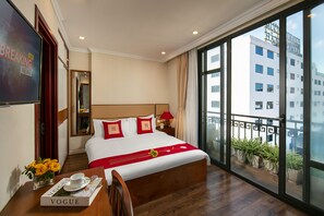 A hotel room with a large bed, a desk with a magazine, a vase with flowers, and a view of buildings through the window. - Au Coeur d'Hanoi Apartment (Hanoi)