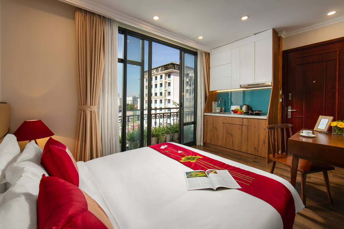 Une chambre d’hôtel dotée d’un grand lit, d’une petite kitchenette et offrant une vue sur la ville grâce à de grandes fenêtres du sol au plafond.