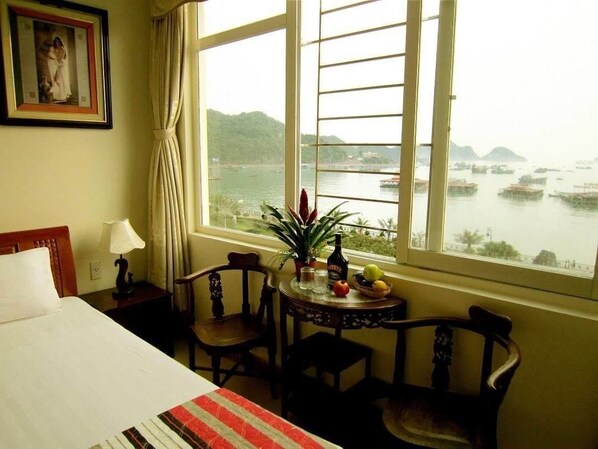 A hotel room with a bed, a desk with a lamp, a chair, a table with a plant and fruit, and a window with a view of boats and hills. - Catba Dream Hotel (Hai Phong)