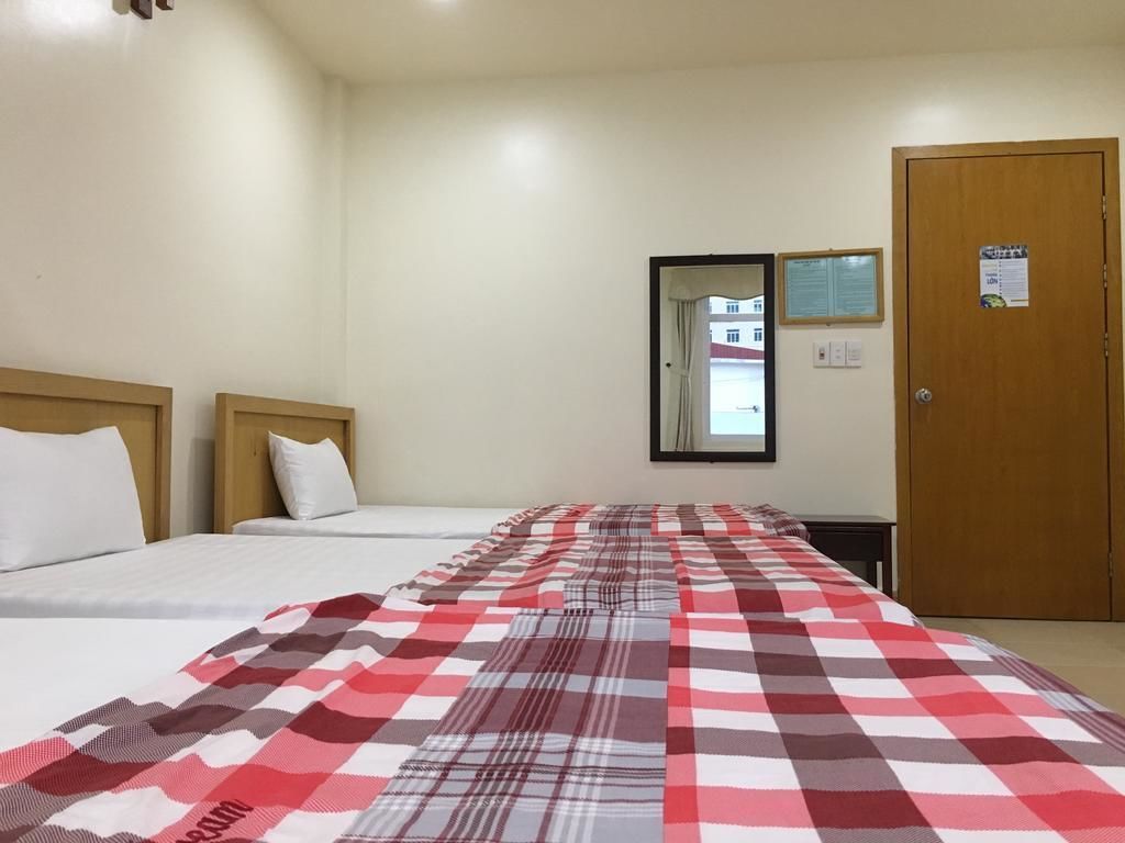 Habitació d'hotel amb dos llits, una manta a quadres, una porta de fusta i una finestra amb vista a edificis.
