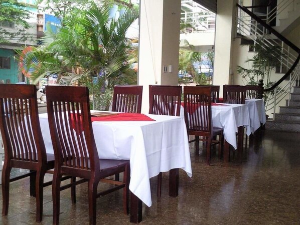 A row of wooden tables with white tablecloths and red placemats, arranged in a covered outdoor area with a staircase in the background. - Catba Dream Hotel (Hai Phong)