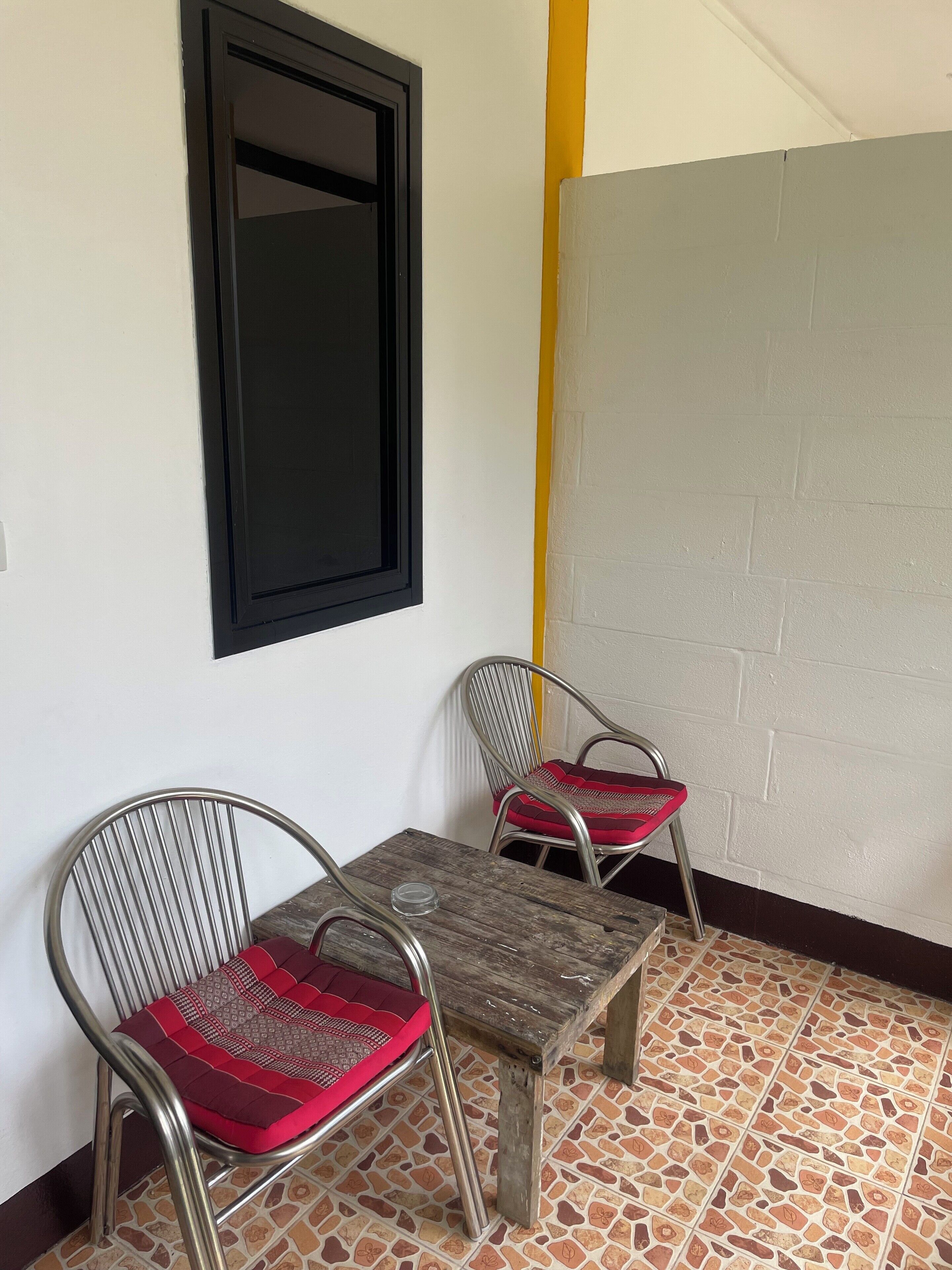 Una habitación pequeña con dos sillas de metal con cojines rojos, una mesa de madera, una ventana con marco negro y un suelo de baldosas.