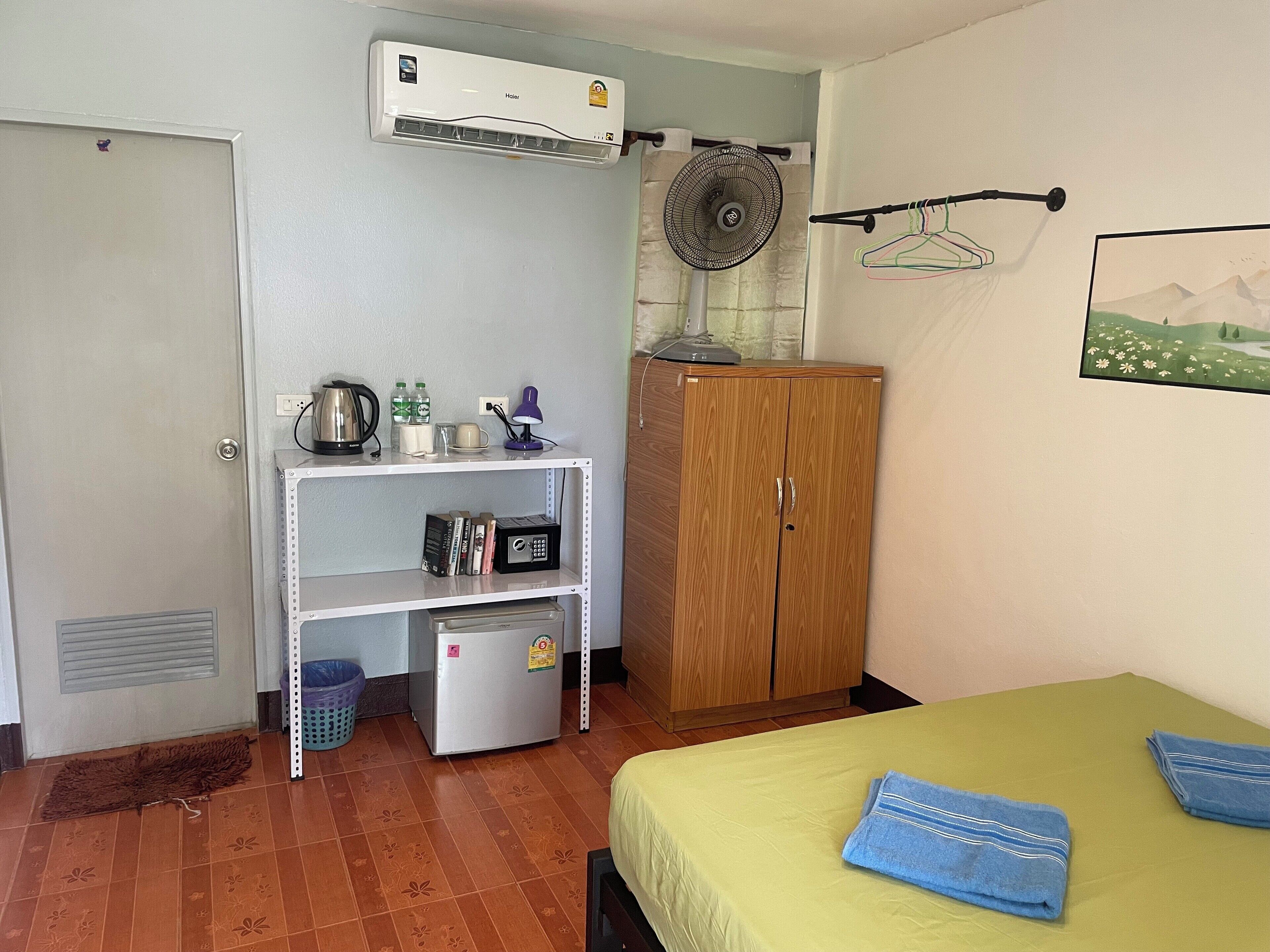 A room with a bed, a wardrobe, a shelf with various items, a fan, and an air conditioner.